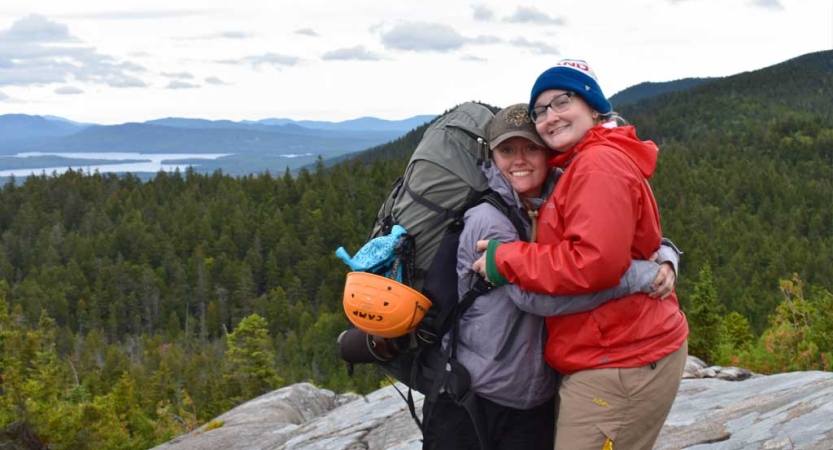 Two people embrace on a rocky overlook above a mountain landscape